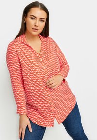 Camicetta rossa con bottoni, caratterizzata da un motivo geometrico a diamante bianco, maniche lunghe e orlo curvo, abbinata a jeans scuri.