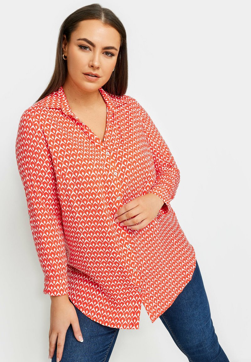Camicetta rossa con bottoni, caratterizzata da un motivo geometrico a diamante bianco, maniche lunghe e orlo curvo, abbinata a jeans scuri.