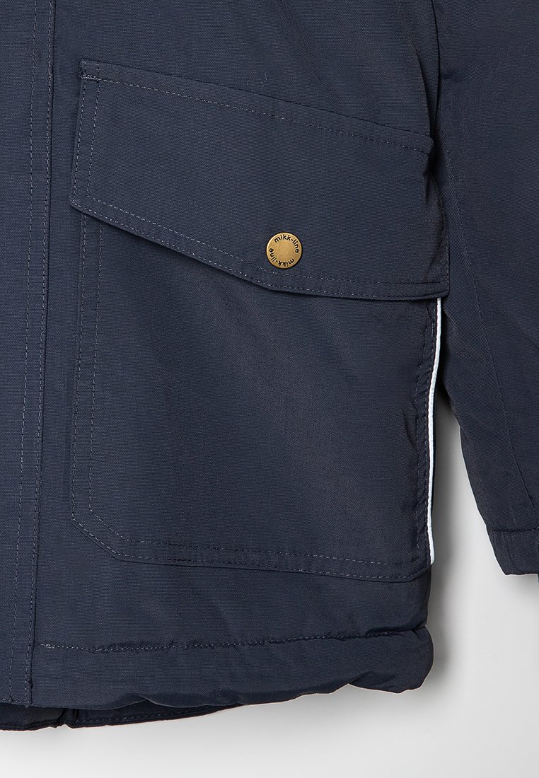 Giacca isolante blu navy con una grande tasca frontale, dettagli in bottone bronzo e accenti bianchi sui lati. Realizzata in tessuto resistente e texture.