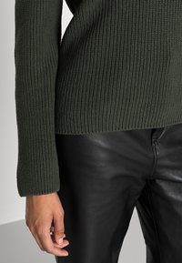 Lähikuva henkilöstä, joka on pukeutunut tummanvihreään ribbineulepaitaan ja mustiin nahkahousuihin, vasen käsi rentona sivulla.