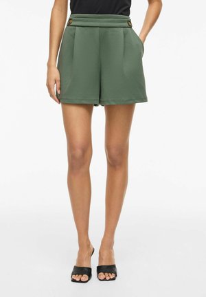 Femeie care poartă pantaloni scurți verde măsliniu cu talie înaltă și nasturi în talie, top negru și pantofi negri cu vârf deschis, stând cu mâinile în buzunare.