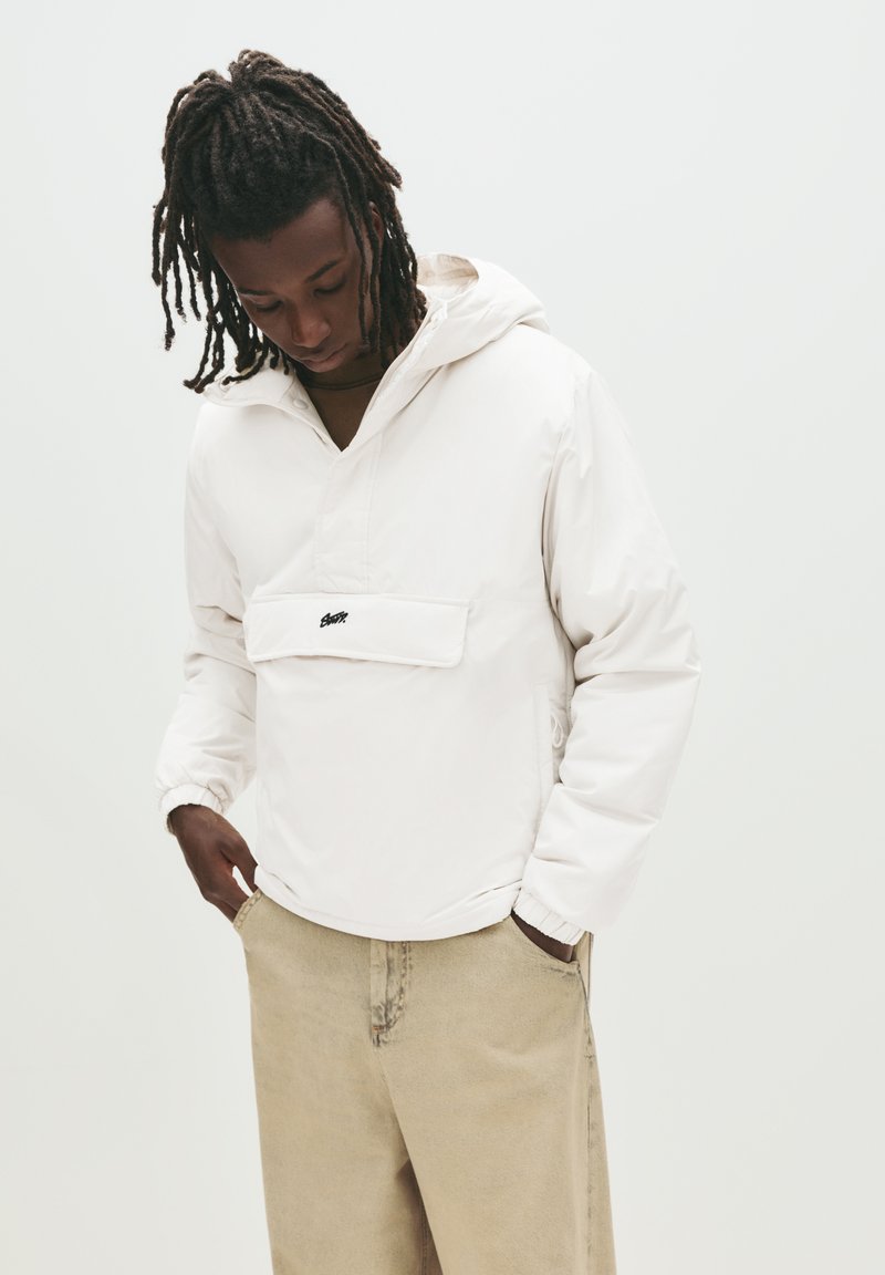 Junge Person mit Dreadlocks, die einen weißen Hoodie und beige Hosen trägt, schaut nach unten mit den Händen in den Taschen vor einem einfarbigen Hintergrund.