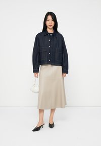 Kvinne som står iført en mørkeblå denimjakke, beige midiskjørt, svarte høyhælte mules og holder en liten hvit veske med rynker.