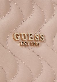 Materiale trapuntato rosa chiaro con cuciture ondulate e un logo dorato "GUESS EST 1981" riportato in modo prominente al centro.