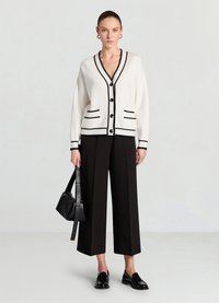 Cardigan crème avec bordure noire, encolure en V, quatre boutons et poches ; associé à un pantalon noir à jambes larges et des mocassins noirs. Petit sac à main inclus.