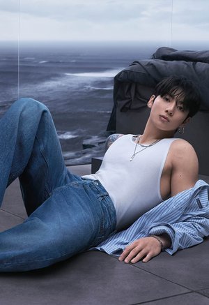 Mladić s tamnom kosom leži na podu blizu oceana, nosi bijelu majicu bez rukava, plave traperice, prugastu košulju prekriženu preko ruke, s nakitom i piercingom.