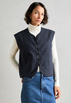 Laivastonsininen klassikkopinnalla varustettu ilman hihoja oleva liivi, jossa on pyöreä pääntie, nappiyksityiskohta ja kaksi etutaskua. Liivi on yhdistetty vaaleancremestä turtleneckista ja farkkuhameesta.