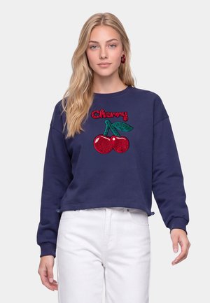 Jonge vrouw met lang blond haar draagt een marineblauwe sweatshirt met rode pailletten kersen en de tekst "Cherry", gecombineerd met een witte broek.
