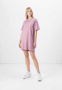 Woman standing in loose mauve T-shirt dress and white high-top sneakers against plain white background.
