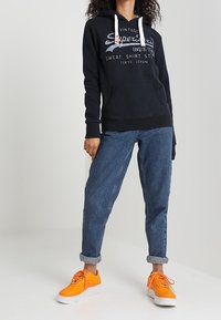 Sudadera negra con cordones blancos, detalle del logo, bolsillo canguro; combinado con jeans azules arremangados y zapatillas brillantes naranjas.