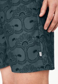 Pánské šortky v tmavě zelené látce se vzorovaným designem obsahujícím spirály a květinové motivy. Obsahují malou značkovou etiketu na bočním švu.