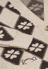 Cardigan lavorato a maglia beige con motivi quadrati marrone scuro che presentano quadrifogli e una parte di volto di gatto, con bottoni di legno visibili.