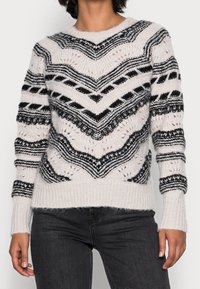 Femme portant un pull blanc en tricot avec des rayures géométriques noires et des motifs, assorti à un jean noir, sur un fond uni.