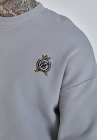Hanorac gri dintr-un material moale, cu guler ribbit și un logo auriu brodat, cu detalii de coroană și laur.