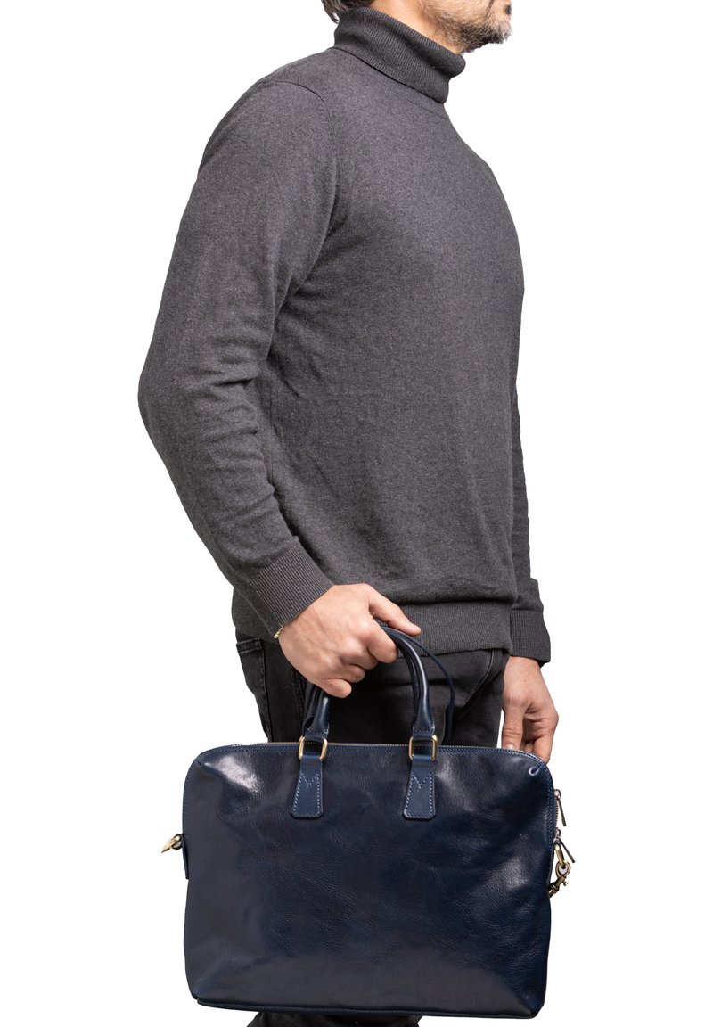 Borsa in pelle color blu navy con due manici superiori, texture liscia, chiusura con zip e dettagli in metallo dorato, tenuta da una persona che indossa un doppiopetto grigio.