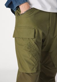 Olīvu zaļas kravas bikses ar gludu tekstūru, ar sānu kabatām, pastiprinātām šuvēm un leņķveida šuvju detaļām.