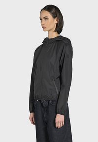 Giacca leggera nera con cappuccio, chiusura a zip frontale, polsini elasticizzati e tasche laterali; realizzata in tessuto liscio e idrorepellente.