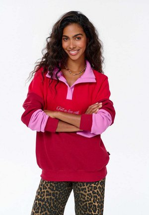 Kobieta z kręconymi włosami uśmiecha się, ubrana w czerwono-różowy sweter, z założonymi rękoma i spodniami w panterkę, na tle jednolitego białego tła.