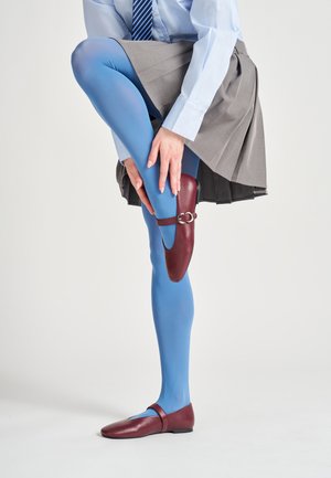 Scarpa di pelle bordeaux con dettaglio a cinturino, abbinata a collant blu e una gonna grigia a pieghe, che mostra una gamba piegata in una postura eretta.