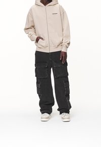 Beige zip-up-hoodie med framfickor, kombinerad med lösa svarta cargobyxor med stora sidofickor. Sneakers fulländar outfiten.