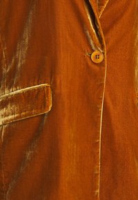 Veste en velours de couleur rouille riche, avec une fermeture à un bouton, des revers crantés et une poche avant au toucher doux.