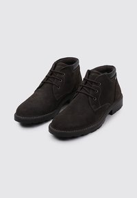Botines de ante marrón oscuro con cuellos acolchados negros, puntas redondas y cierres de cordones negros sobre un fondo gris claro.