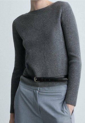 Femme portant un pull gris côtelé ajusté rentré dans un pantalon gris clair avec une ceinture noire, main dans la poche, sur un fond uni.