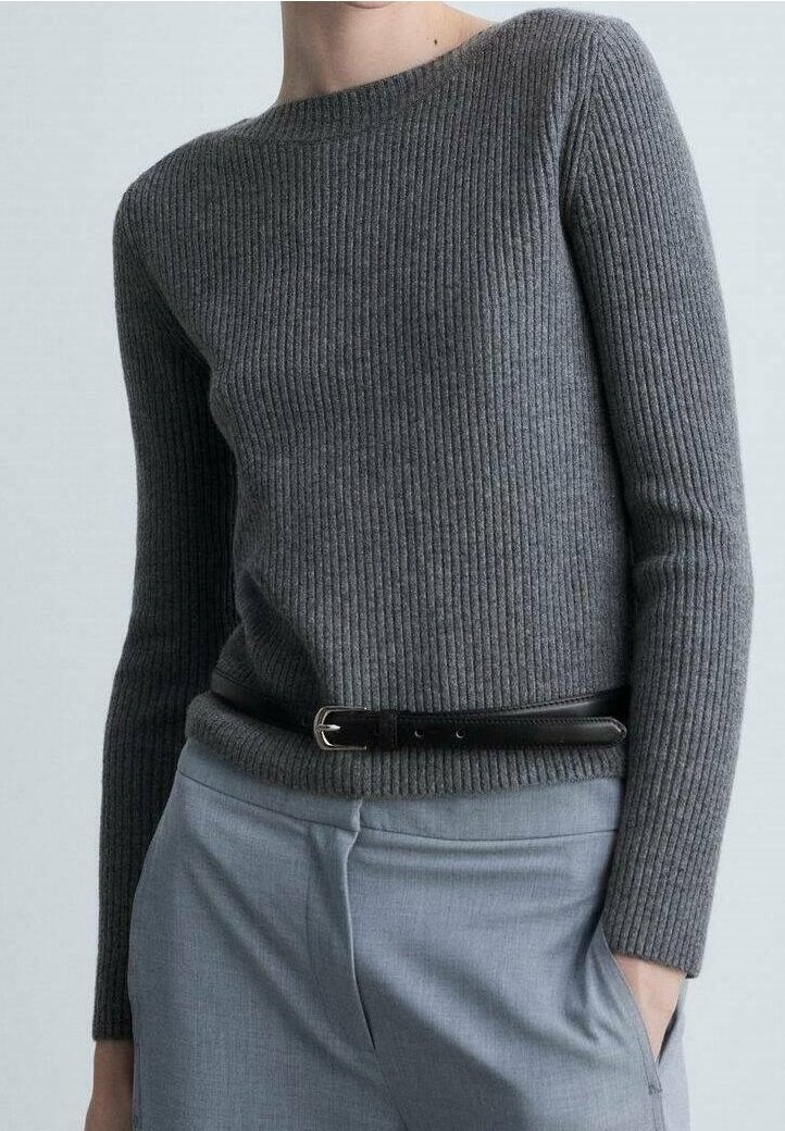 Femme portant un pull gris côtelé ajusté rentré dans un pantalon gris clair avec une ceinture noire, main dans la poche, sur un fond uni.