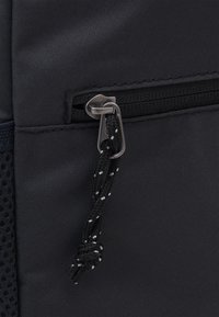 Borsa in tessuto nero con tasca con zip, dotata di un tirante in metallo argentato e di un cordino nero con accenti bianchi. Superficie testurizzata, materiale resistente.