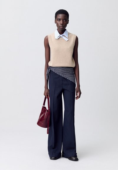 Persona alta che indossa un maglione senza maniche beige sopra un colletto di camicia a righe, pantaloni a gamba larga navy con dettaglio avvolgente, mentre tiene una borsa bordeaux.