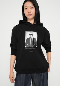 Fekete kapucnis pulóver, puha anyagból, elöl egy napszemüveges férfi grafikájával és öltönnyel; zsinórral állítható kapucnival és bordázott mandzsettákkal rendelkezik.