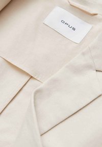 Tissu couleur crème avec une étiquette de vêtement blanche indiquant "OPUS", cousue à l'intérieur d'un vêtement plié, montrant les coutures et les détails de la texture.