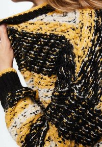 Neulepusero, jossa on rohkeat mustat, keltaiset ja valkoiset värit, teksturoitu kuvio ja ribbireunaiset mustat kaula-aukko ja hihansuut.