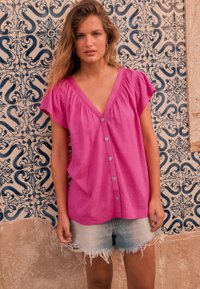 Blouse rose à manches courtes avec boutons, encolure en V et coutures froncées aux épaules, associée à un short en jean effiloché sur un fond à motifs.