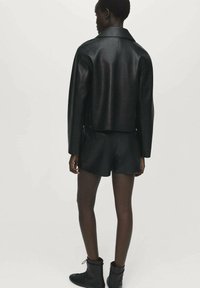 Zwarte leren cropped jack met een strakke silhoutte, brede kraag en gladde textuur. Gecombineerd met zwarte shorts en enkellaarsjes.