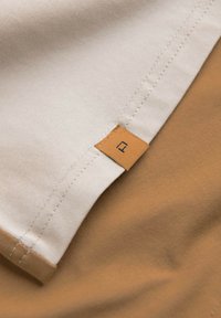 Beige a hnědé vrstvy látky se zašitým lemem. Obsahuje čtvercovou béžovou etiketu s černým symbolem na béžové ploše.