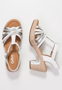 Białe i srebrne sandały z krzyżującymi się paskami, teksturowanym brązowym obcasem na platformie i zapięciem na klamrę z boku. Gładkie, błyszczące materiały.