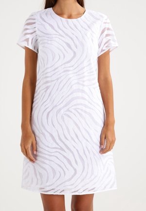 Femme portant une robe blanche à manches courtes, longueur aux genoux, avec un motif texturé à rayures zébrées et un col rond.