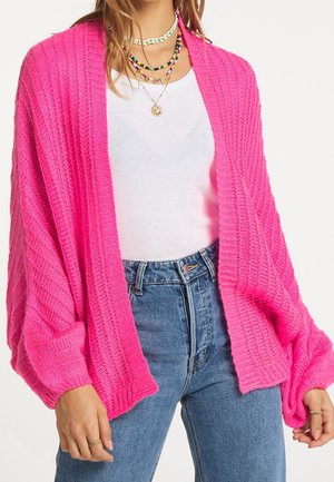 Woman wearing bright pink chunky knit cardigan over white top and blue jeans, accessorized with layered colorful beaded and gold necklaces.