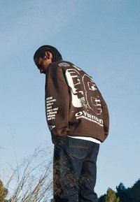 Smeđa oversized dukserica s bijelim grafičkim dizajnom na leđima i rukavu, u kombinaciji s tamnim trapericama. Vanjsko okruženje s plavim nebom.