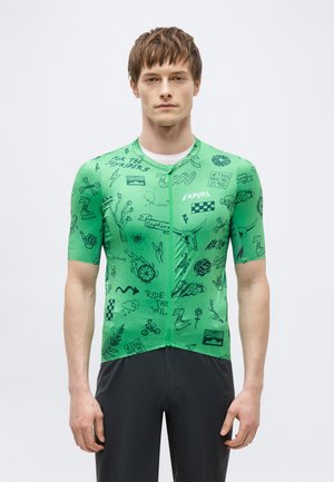 Mann kledd i en grønn sykkeltrøye med korte ermer, dekorert med svarte grafiske krusseduller og tekst, kombinert med svarte bukser, mot en ensfarget bakgrunn.