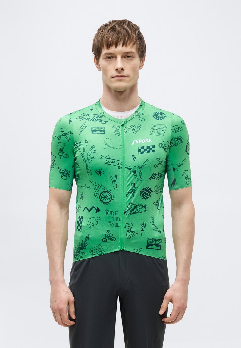 Άνδρας που φοράει πράσινο κοντομάνικο ποδηλατικό μπλουζάκι με μαύρα γραφικά σχέδια και κείμενο, συνδυασμένο με μαύρο παντελόνι, σε απλό φόντο.