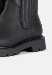 Bota de tobillo de cuero negro con detalle acanalado vertical, suela de goma gruesa y costuras resistentes. Textura suave y diseño de punta redonda.