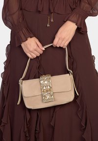 Sac à main en daim beige de forme rectangulaire avec un avant texturé. Il présente une décoration dorée ornée de pierres précieuses claires sur le fermoir.