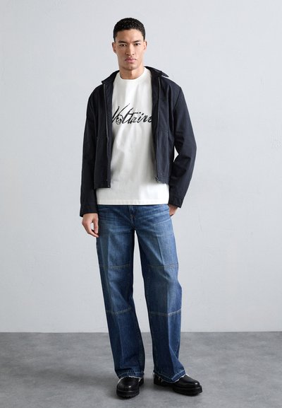 Chaqueta negra con cremallera sobre una camiseta blanca con el texto "Voltair"; jeans azul de corte suelto con costuras en los paneles; zapatos negros de suela gruesa.