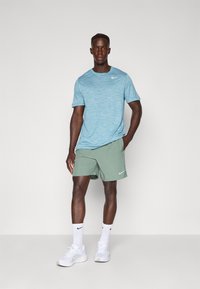 Hellblaues sportliches T-Shirt mit strukturierten Streifen, kombiniert mit grünen Shorts. Weißes Nike-Logo auf beiden. Modell trägt weiße Sneaker und Socken.
