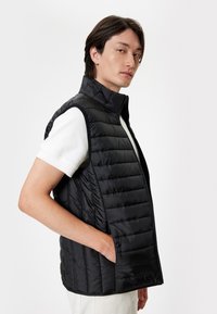 Gilet imbottito nero con colletto alto, design trapuntato e tasche laterali, indossato sopra una camicia bianca. Tessuto liscio e lucido con una vestibilità aderente.