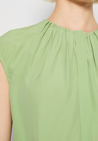 Femme portant un chemisier vert clair sans manches avec des plis froncés autour de l'encolure et un petit détail en encoche.