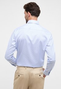 Camicia a righe blu chiaro con colletto button-down e maniche lunghe, abbinata a pantaloni beige con tasche posteriori e dettagli in bottone.