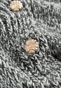 Sivi i bijeli pleteni materijal s teksturiranim uzorkom, s naglaskom na zlatno uvučen dugme. Mekan, topao materijal s vidljivim šavovima.